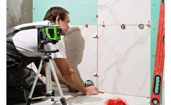 A professional tiler installing large-format marble-look porcelain tiles on a wall using leveling spacers and a laser level for precise alignment.